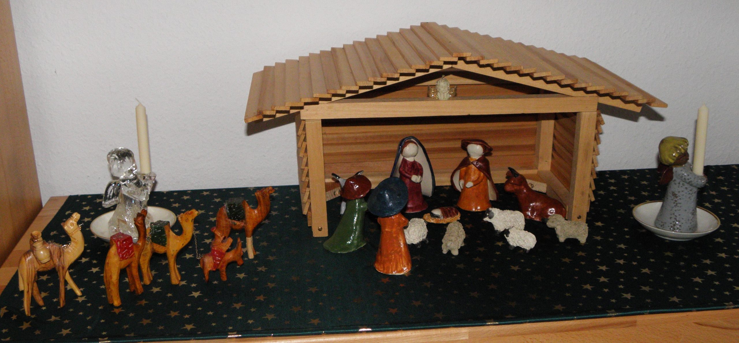 Ein selbstgebastelte Krippe aus Ton. Die Heilige Familie, 2 Hirten und Tiere sind in einem offenen Stall aus Holz. Eine Kamelkarawane nähert sich dem Stall. " Engelfiguren tragen Kerzen.
