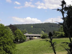 Im Vordergrund befindet sich eine große Bergwiese. Dahinter liegt ein Haus am Waldrand. Im Hintergrund ist der Bocjsberg mit Turm erkannbar
