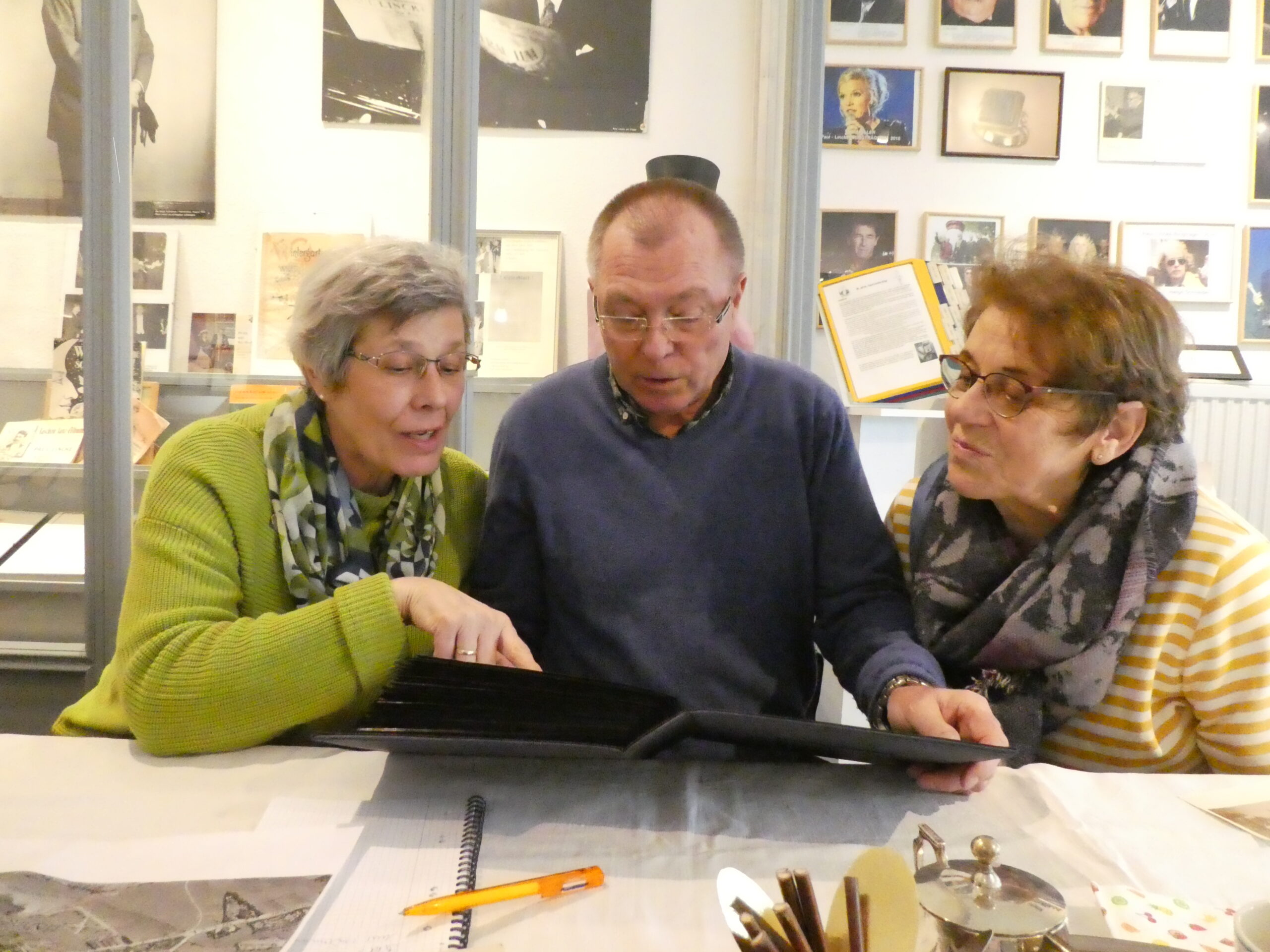 3 Besucher stöbern in einem Fotoalbum. eine Frau (links im Bild) zeigt auf ein Foto. eine anderen Frau (rechts) schaut interessiert auf das Detail.In der Mitte sitzt ein Mann, der das Fotoalbum aufgekklappt hält.