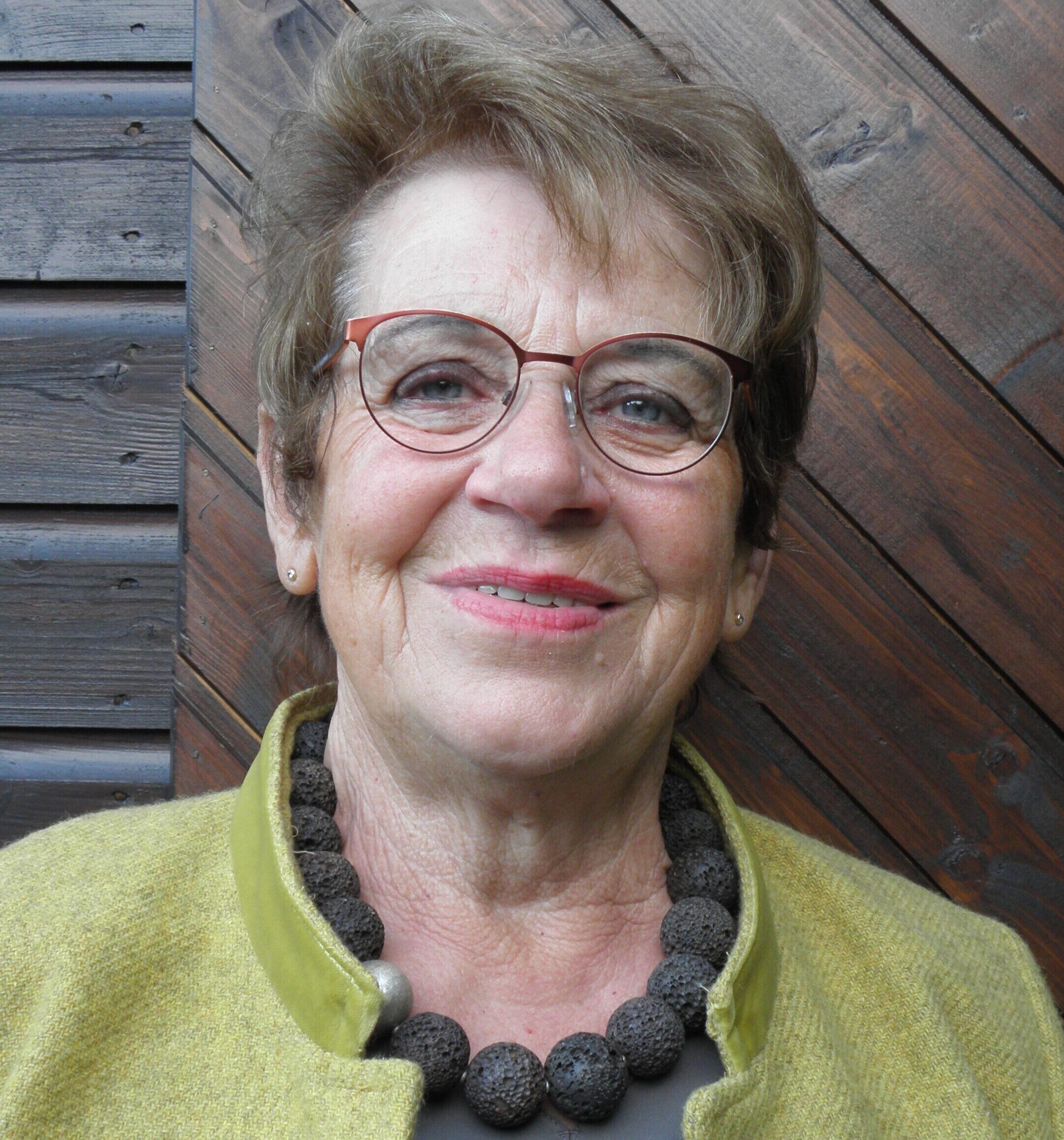 Eine Frau schaut lächelnd in die Kamera. Sie trtägt eine Brille, einen hellgrünen Blouson und hat eine Kette aus grauen , großen Kugeln umgelegt.