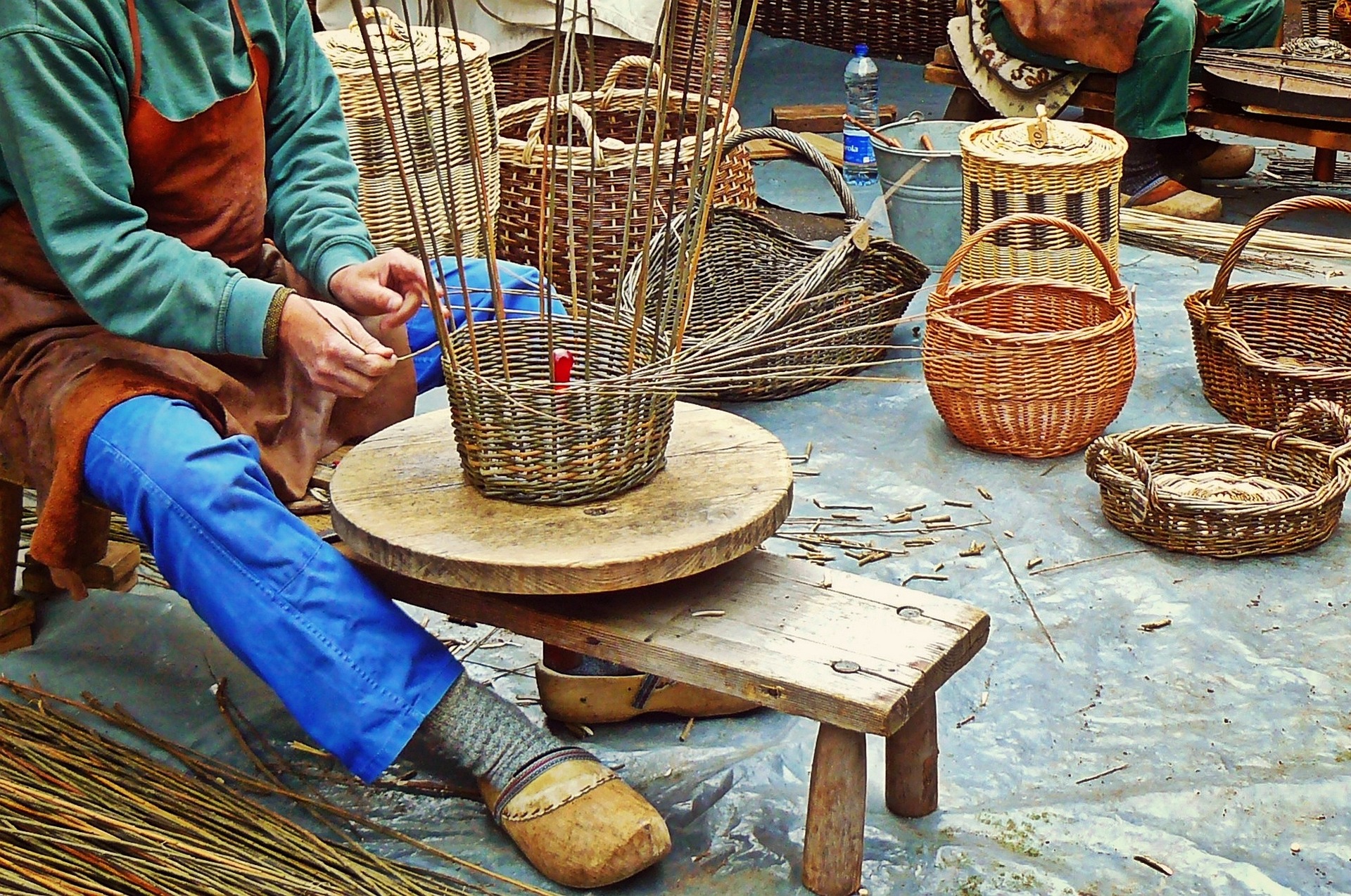 Eine Person sitzt und flicht einen Korb. Im Hintergrund stehen weitere selbstgemachte Körbe aus verschiedenen Naturmaterialien