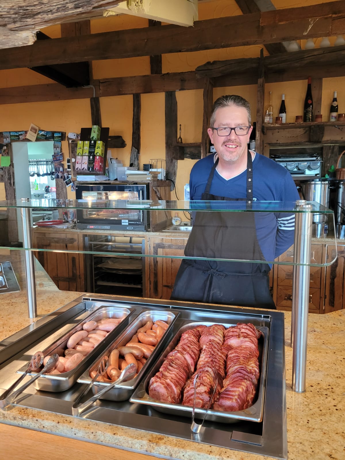 Ein Mann mit einer Schürze steht hinter einem Fleisch- und Wurstbüffet. Er freut sich über seine Gäste: